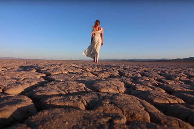 A white woman in a white flowy dress, her back towards the camera, walks along a cracked desert landscape.