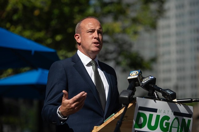 Candidate Matt Dugan wears a dark suit and close-buzzed hair while speaking on the campaign trail.