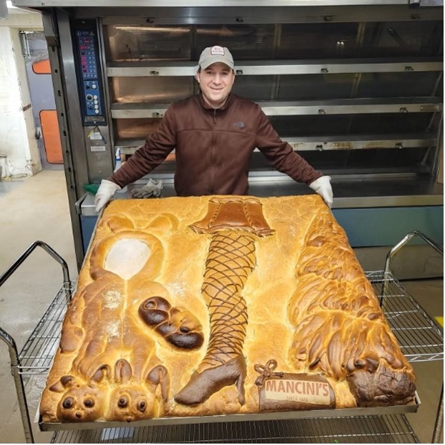 A baker wearing a brown shirt, white gloves, and a white baseball cap, proudly poses with a large bread sculpture