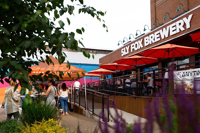 Several groups of people stand and sit at tables outside of a building with a "Sly Fox Brewery" sign