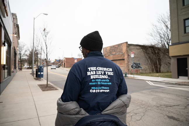 A dark-skinned man in beanie and glasses pulls down his jacket to reveal a shirt with the Bible Center Church's social media accounts on the back
