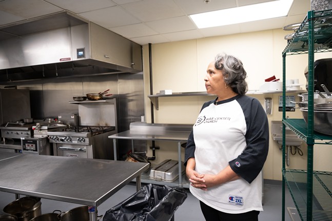 A woman with brown skin and wavy silver hair wearing a baseball tee stands in a clean commercial kitchen.