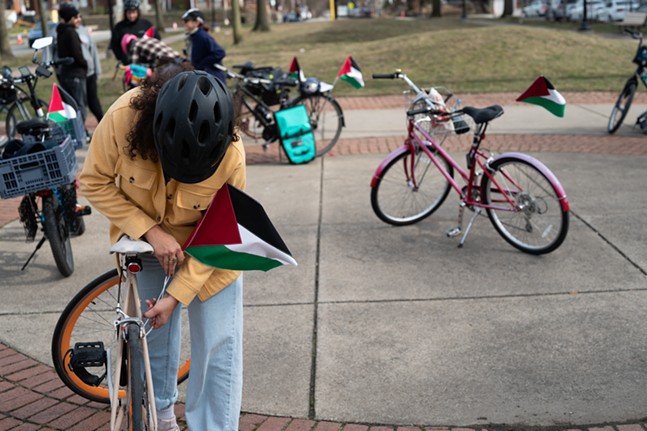 A person with long hair covered by a bike helmet mounts a Palestinian flag to their seatpost tube
