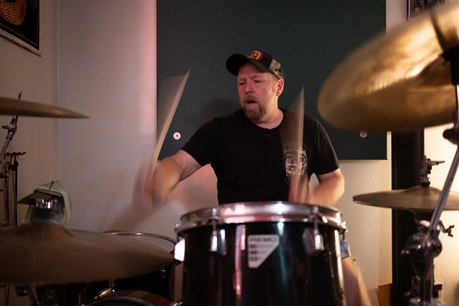 A drummer with a salt-and-pepper goatee and trucker hat has blurry hands in a shot of him pounding the toms
