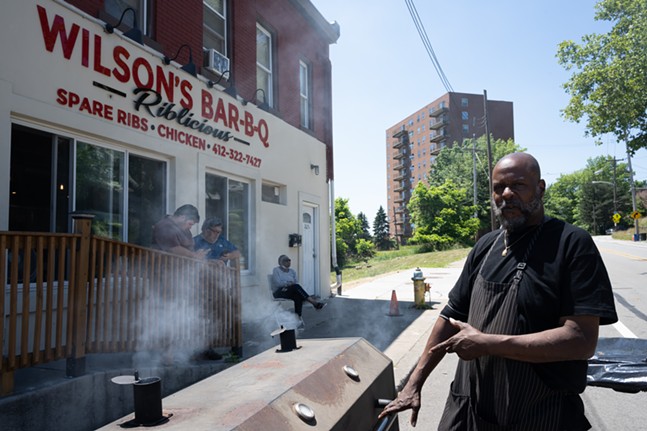 A man points to a large smoker as hungry diners lean on a railing while waiting for their order