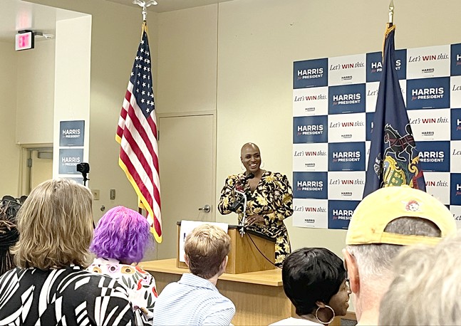A smiling woman wearing a patterned dress and shaved head at a room full of Harris for President signs