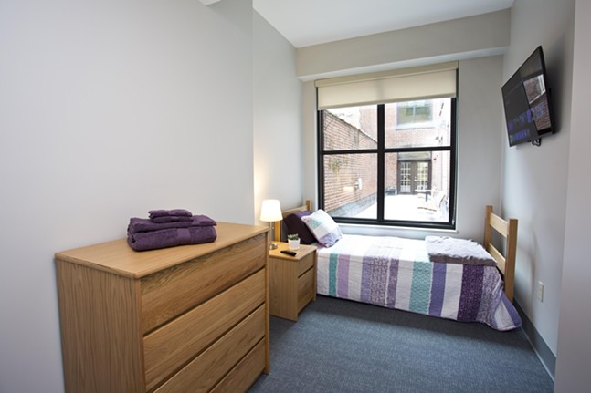 A spacious bedroom with dorm-style wooden furniture and a wall-mounted TV