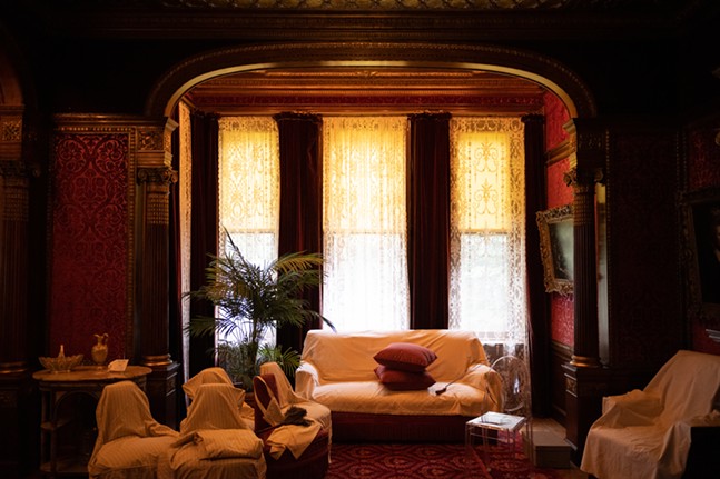 Furniture sits covered in a room lined with ornate woodcarving and silk wall coverings