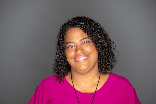 A woman with brown skin and tightly curled hair down to her shoulders wears a pink shirt and necklace