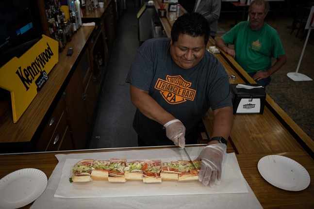 A man smiles as he cuts a two-foot-long sandwich into three-inch pieces on a wooden counter.