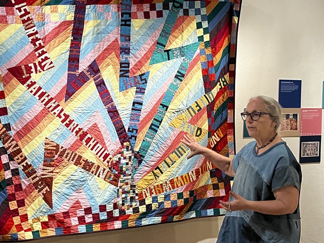 A woman in a collaged denim dress and glasses stands near a quilt of neon lightning bolts