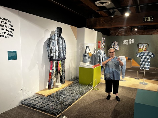 A woman with grey hair and a collaged denim dress stands near a figure made of scrap fabric