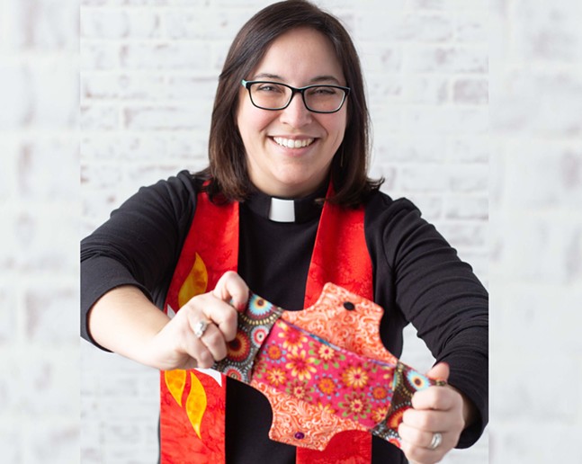 A woman with shoulder-length brown hair and glasses in a pastor's frock and color holding a red patterned fabric maxi pad