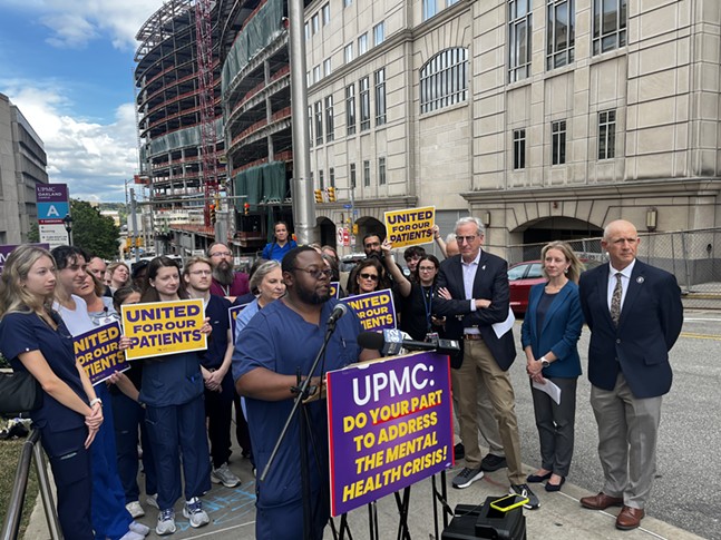 A nurse with short hair, a beard and glasses speaks before a crowd in scrubs and suits near large hospital buildings