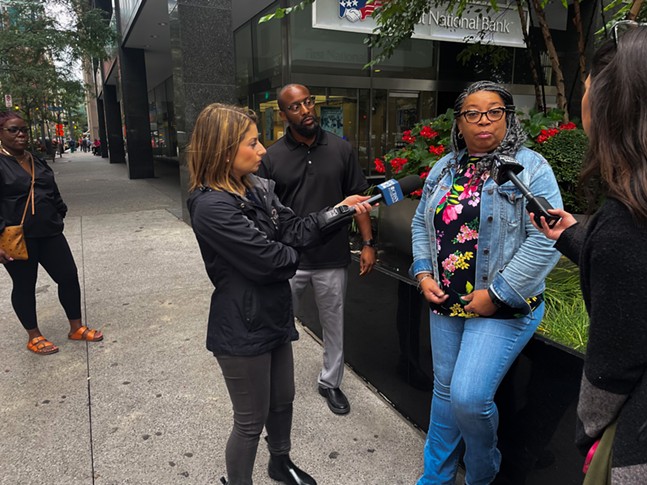 A woman in glasses in a denim jacket and pants speaks with two television reporters holding microphones