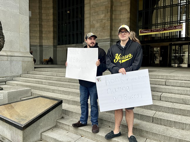 Two young light-skinned men in Pittsburgh sports apparel carry homemade signs urging Bob Nutting to sell the Pirates