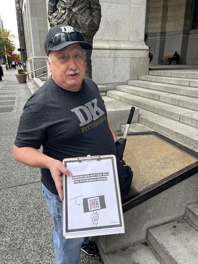 An older man with a mustache holds a clipboard with a QR code for a petiton