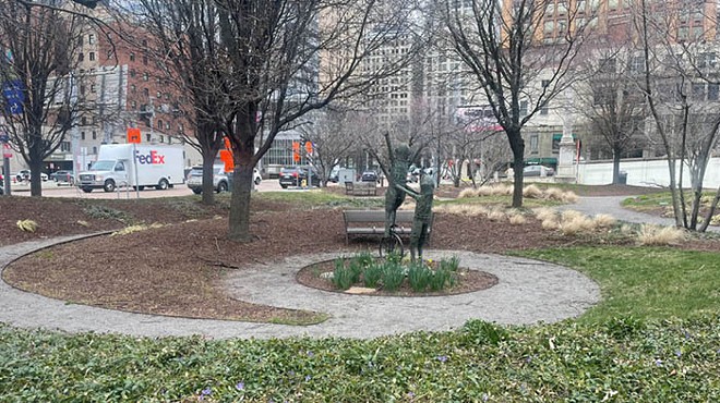 Circular path goes around a sculpture, surrounded by trees and small purple flowers