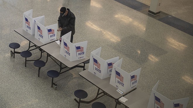 A man in a jacket and cap votes at a folding table in a large school cafeteria.