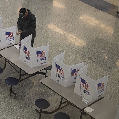 A man in a jacket and cap votes at a folding table in a large school cafeteria.